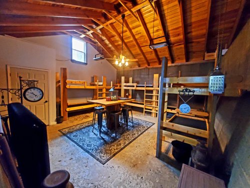 A spacious room with wooden shelves, a table with stools, and a high ceiling featuring exposed beams and a ceiling fan.