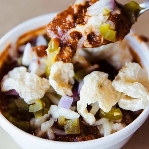 A bowl of chili topped with onions, jalapeños, cheese, and crispy pork rinds, with a spoonful lifted above.