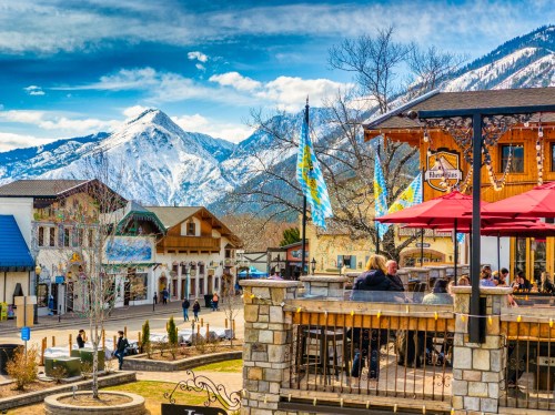 A scenic view of a mountain town with outdoor dining, colorful flags, and snow-capped mountains in the background.