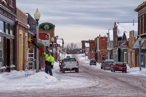 A snowy street scene with a worker hanging holiday decorations on a building, while a truck is parked nearby.