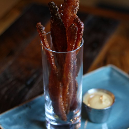 A glass filled with crispy bacon strips, served alongside a small candle on a blue plate.