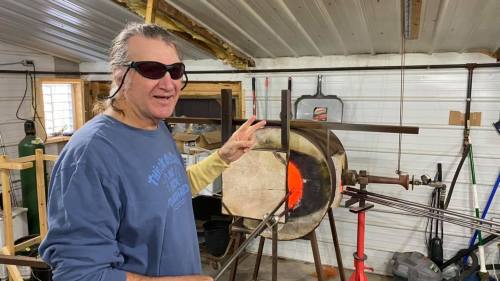 A man in sunglasses stands next to a glassblowing furnace, holding a tool and smiling in a workshop setting.