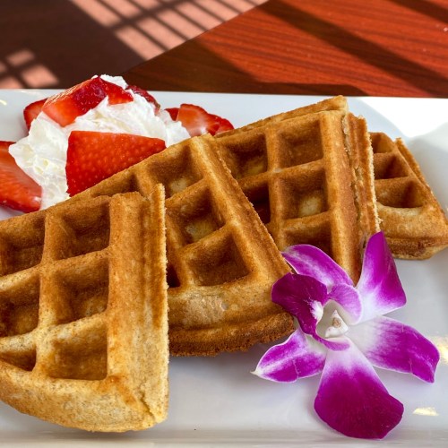 A plate of golden waffles served with strawberries, whipped cream, and a purple orchid garnish.