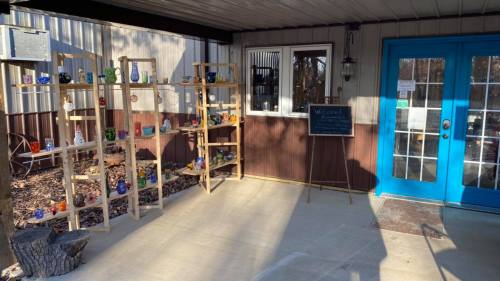 A sunny entrance with blue doors, wooden shelves displaying colorful glass vases, and a chalkboard sign.