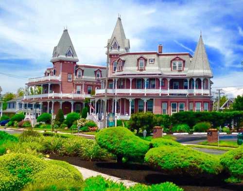 Victorian-style mansion with multiple towers, surrounded by lush greenery and a clear blue sky.