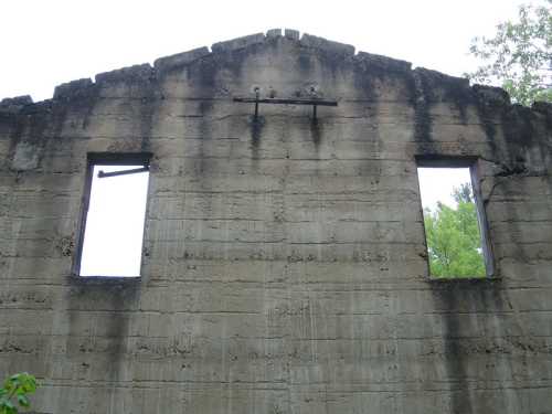 A weathered concrete wall with two empty windows, surrounded by greenery and a cloudy sky.