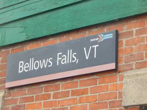 Sign for Bellows Falls, VT, featuring Amtrak logo, mounted on a brick wall with green wooden trim.