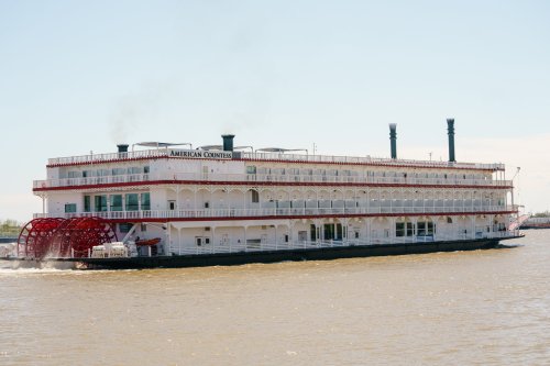A large riverboat with a red paddle wheel sails on a calm river under a clear blue sky.