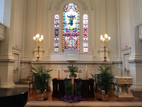 A church altar with a wooden cross, candles, and plants, framed by a colorful stained glass window.