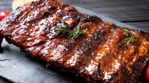 Close-up of glazed, grilled ribs garnished with herbs, served on a dark slate platter with cherry tomatoes.
