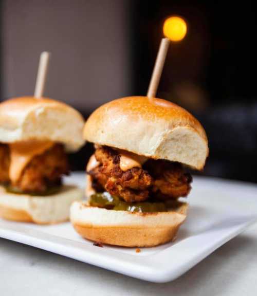 Two mini chicken sandwiches on a white plate, topped with pickles and sauce, served with toothpicks.