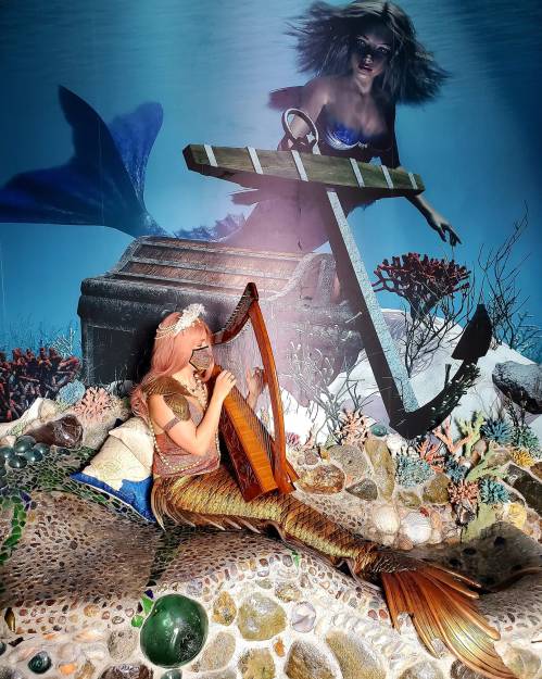 A mermaid playing a harp on a rocky ocean floor, with a mysterious figure swimming in the background.