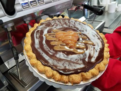 A large pie with a glossy chocolate topping and caramel drizzle, held near a coffee machine.
