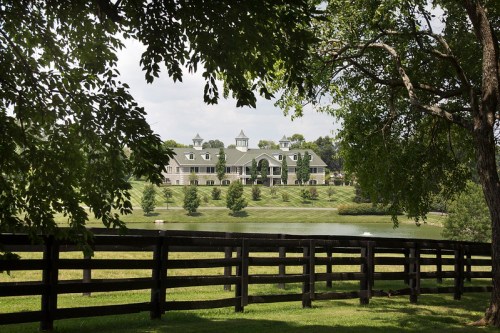 A large, elegant building surrounded by greenery and a pond, framed by trees and a wooden fence.