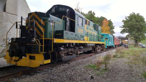 A green and yellow locomotive with a train of colorful cars on a gravel track, surrounded by trees and a bridge.