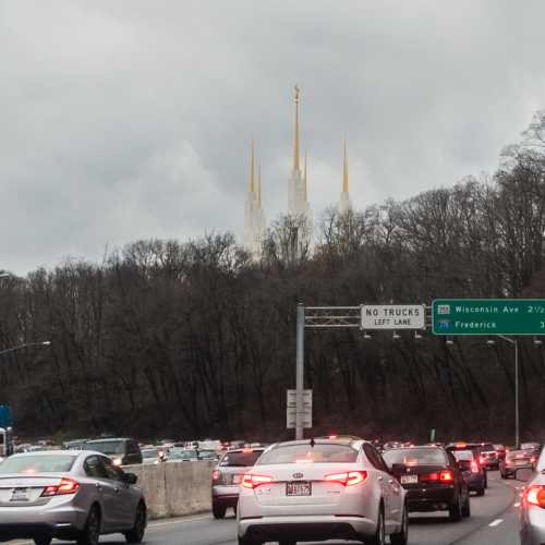 Traffic on a highway with a distant view of tall spires and a cloudy sky. Road signs indicate directions ahead.