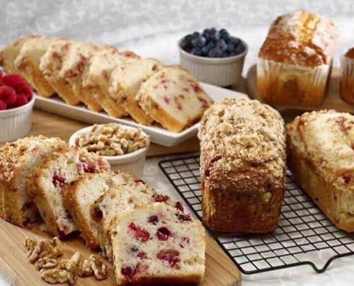 Sliced fruit bread with raspberries and blueberries, displayed on wooden boards with whole loaves and nuts.