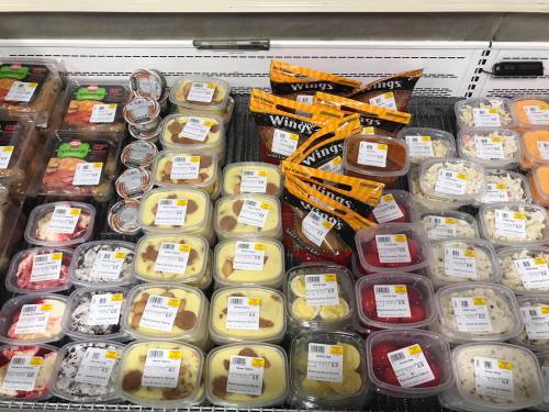 A display of various packaged food items, including desserts and chicken wings, with price tags visible.