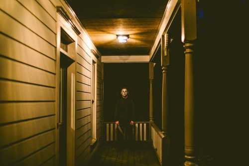 A figure in a mask stands in a dimly lit porch, holding a knife, creating a suspenseful and eerie atmosphere.