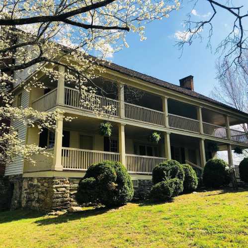 A charming two-story house with a wraparound porch, surrounded by greenery and blooming trees under a clear blue sky.