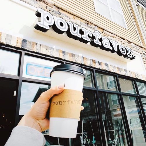 A hand holding a coffee cup with a sleeve in front of a café named "pourfavor." Windows and an open sign are visible.