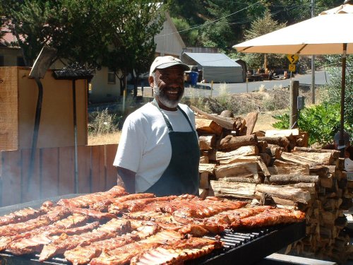 A smiling man stands by a grill filled with ribs, surrounded by firewood and a sunny outdoor setting.