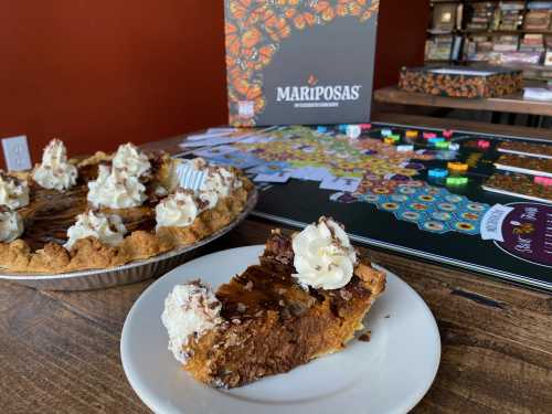 A slice of pie with whipped cream on a plate, next to a whole pie and a colorful board game in a cozy setting.