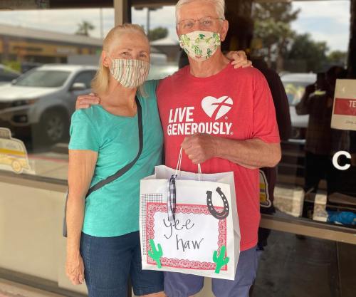 A smiling couple wearing masks stands together outside a store, holding a gift bag that says "Yee Haw."