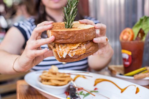 A person holds a stacked sandwich with a sprig of rosemary, accompanied by fries and a drink on a table.