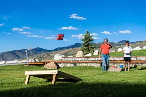 A man and a boy play cornhole outdoors, with mountains and a blue sky in the background. A red bean bag is in the air.