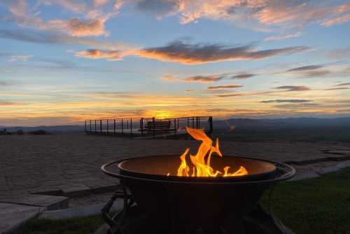 A fire pit with flames in the foreground, set against a colorful sunset and distant mountains.