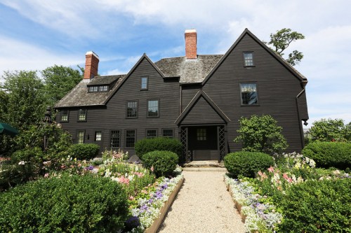 A large black wooden house with multiple gables, surrounded by lush greenery and colorful flower beds.