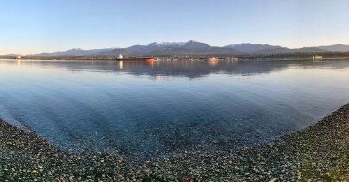 A serene waterfront view with calm water, distant mountains, and ships on the horizon under a clear sky.