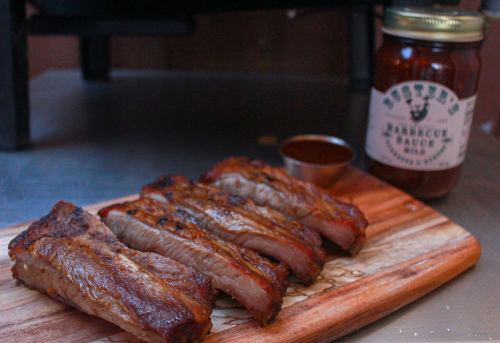 Grilled ribs on a wooden board with a small bowl of sauce and a jar of barbecue sauce in the background.