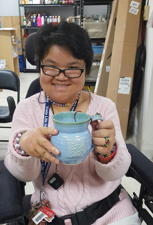 A smiling person in a pink sweater holds a decorative blue pottery mug, sitting in a workshop environment.