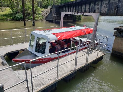 A jet boat with a red canopy is docked at a pier, ready for rides on a calm river surrounded by greenery.