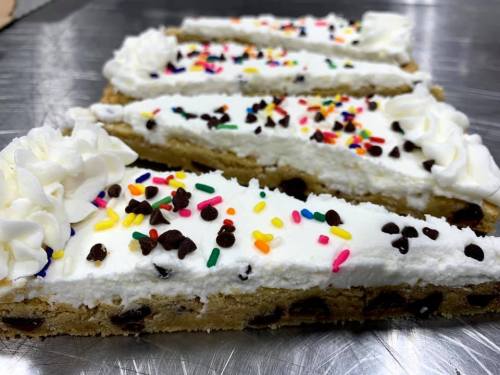 Three slices of cookie cake topped with whipped cream, chocolate chips, and colorful sprinkles on a silver surface.