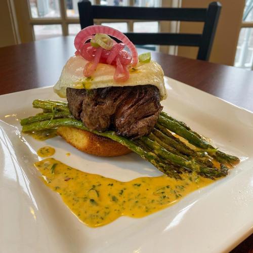 A gourmet dish featuring a beef patty topped with a fried egg, asparagus, pickled onions, and a yellow sauce on a plate.