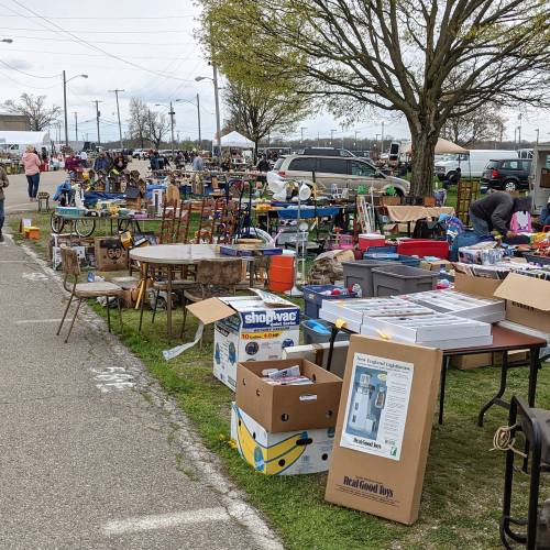 A bustling outdoor flea market with various items for sale, including furniture, tools, and toys, set up along a grassy area.