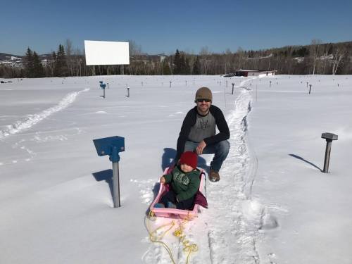 A man kneels beside a child in a sled on a snowy landscape, with trees and a clear blue sky in the background.