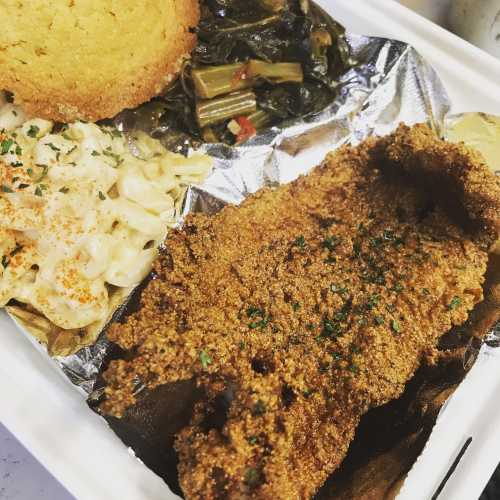 A plate featuring fried fish, macaroni and cheese, collard greens, and cornbread, all served in a takeout container.