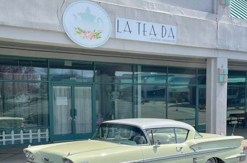 A vintage car parked in front of a tea shop named "La Tea Da," featuring a charming storefront.