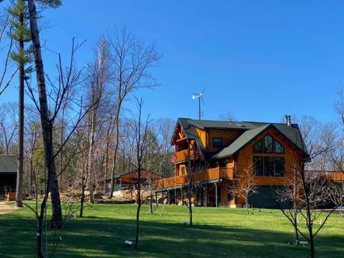 A large wooden house with a deck, surrounded by trees and a clear blue sky, featuring a small wind turbine on the roof.