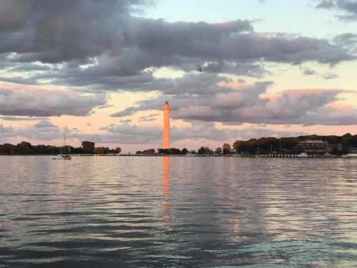 A lighthouse stands tall by the water, reflecting light under a colorful sky with clouds at sunset.