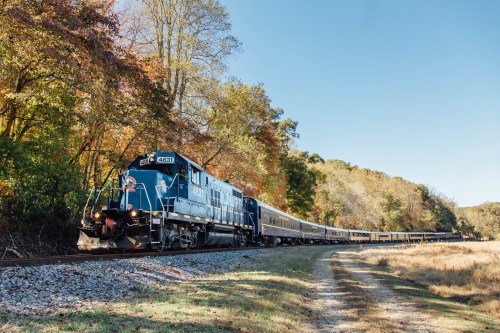A blue train travels along a scenic route surrounded by trees with autumn foliage under a clear sky.