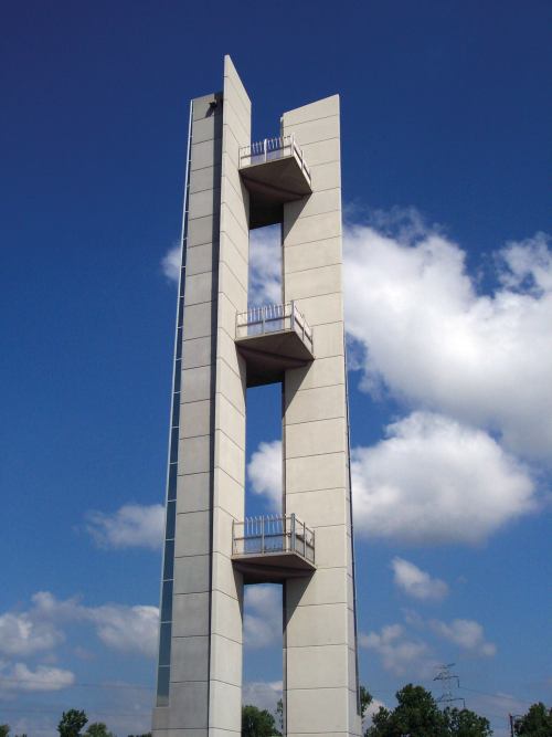 A tall, modern concrete tower with open sections and balconies, set against a blue sky with fluffy white clouds.