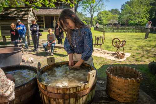 A girl washes clothes in wooden tubs while children watch in a historical outdoor setting.