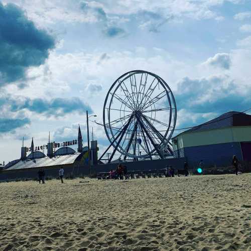 A beach scene featuring a large Ferris wheel against a cloudy sky, with people and structures in the background.