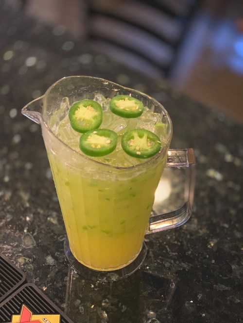 A pitcher of yellow-green drink with ice and sliced jalapeños on top, placed on a dark countertop.