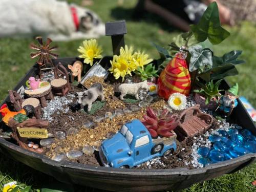 A colorful miniature garden scene with figurines, flowers, and a blue toy truck in a decorative planter.